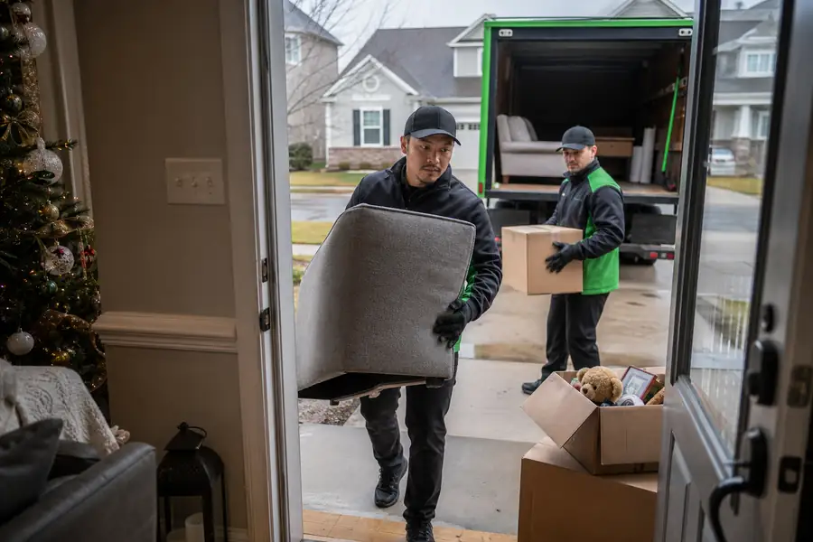 Green-accented junk removal truck with uniformed two-person crew, festive living room being tidied with donation boxes by the door