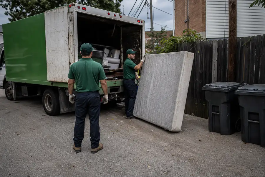 Green-accented junk removal truck with uniformed two-person crew, box spring leaning next to trash bins in an alley