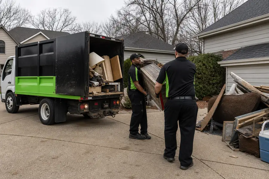 Green-accented junk removal truck with uniformed two-person crew, junk removal truck being loaded with household junk in a suburban driveway