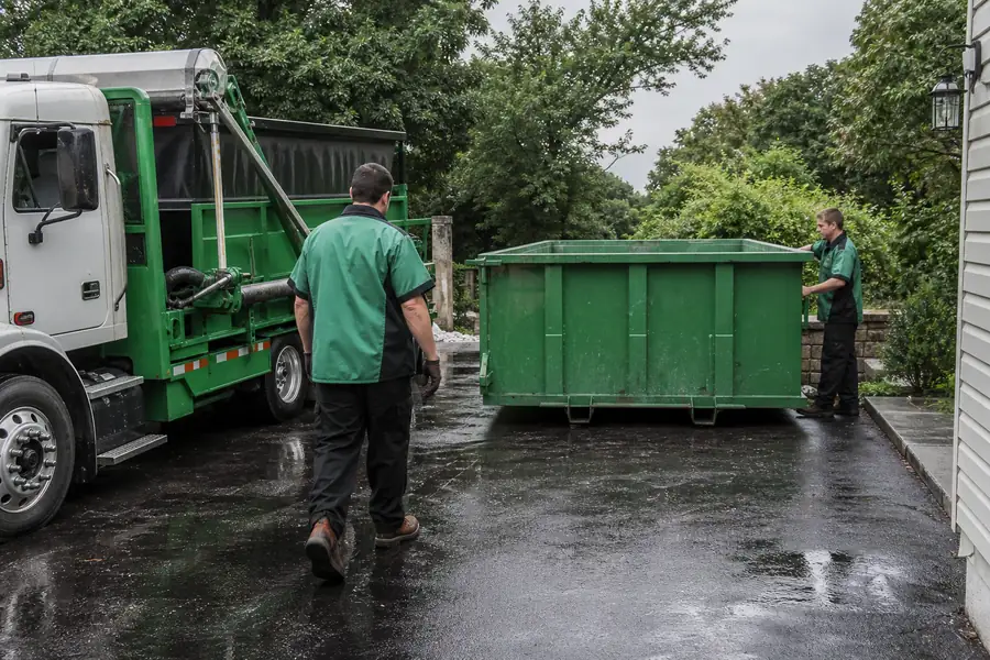 Green-accented junk removal truck with uniformed two-person crew, roll-off dumpster in a driveway next to a junk removal truck