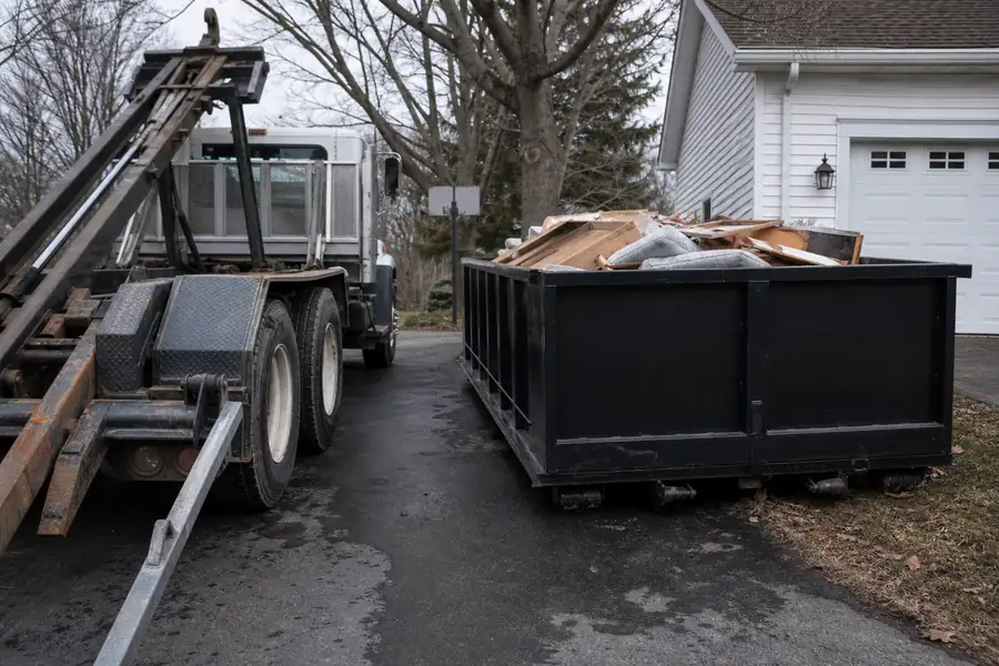 Roll-off dumpster in a driveway next to a junk removal truck