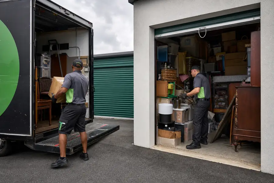 Green-accented junk removal truck with uniformed two-person crew, open self-storage unit packed floor to ceiling with boxes and furniture